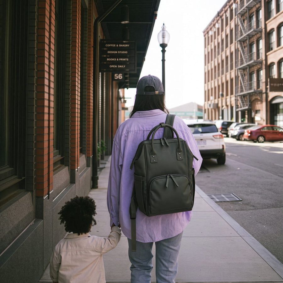 Parent walking a toddler down a city street, wearing the olive green ALUXO Active X baby bag backpack