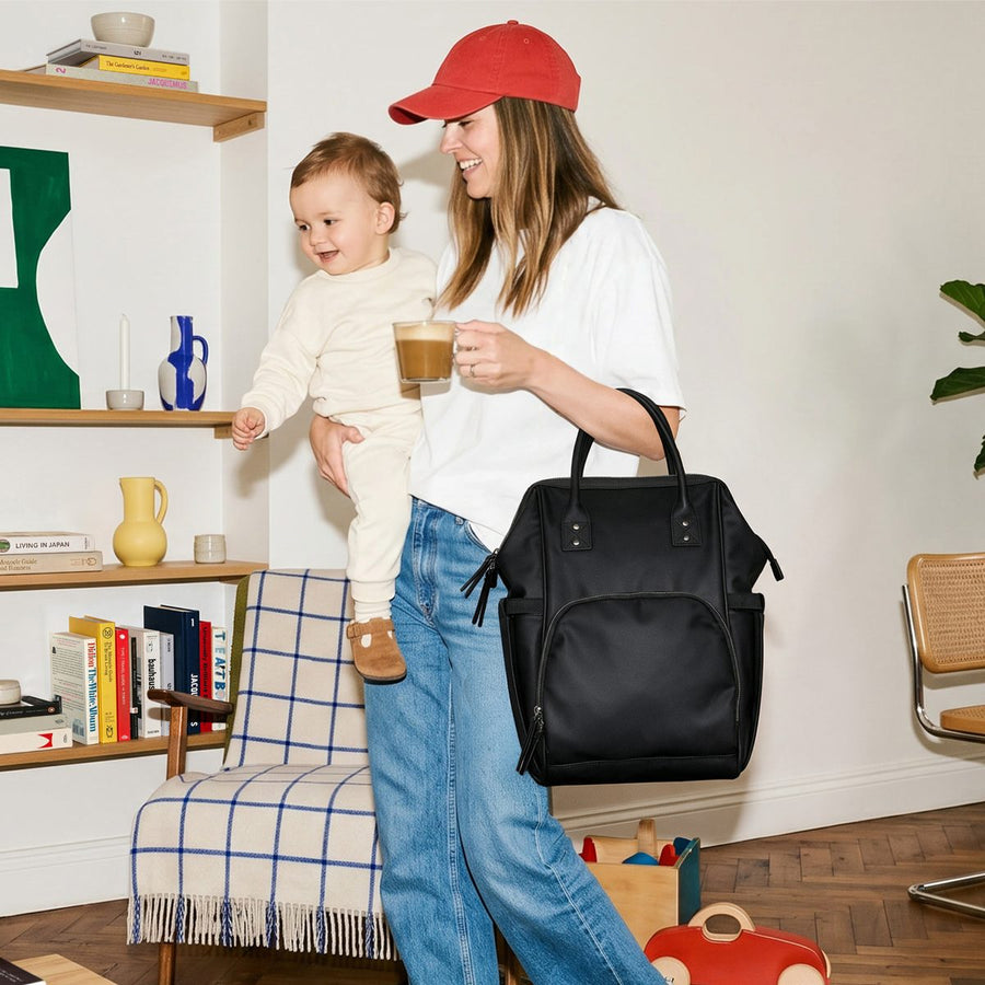 Mum in a red cap carrying her baby and a coffee, with the black ALUXO Active X baby bag on her shoulder
